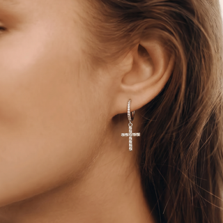 A close-up of a woman's ear wearing a delicate silver cross dangle earring adorned with small clear stones.