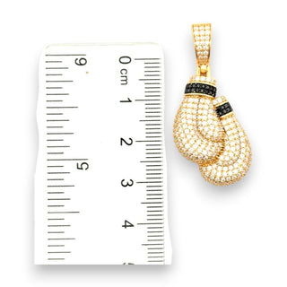 Gold boxing gloves pendant paved with clear stones, with black gemstone accents, shown next to a white ruler for scale.