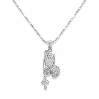 A silver-toned box chain necklace featuring a praying hands pendant paved with small clear stones, with a dangling cross charm, shown on a plain white background.