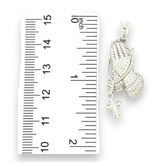 A silver praying hands pendant with a small cross charm, shown next to a white inch ruler for scale on a white background.