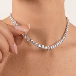 A close-up of a model's neck and hand wearing a silver tennis necklace with clear round-cut stones, set against a plain light background.