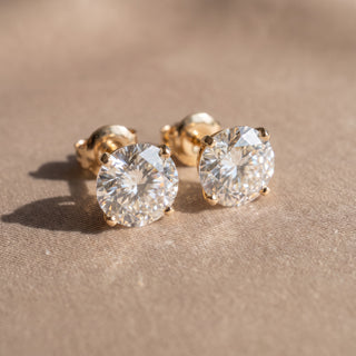 Superior Silver Portuguese cut moissanite stud earrings in gold vermeil with four-prong settings on a beige surface.